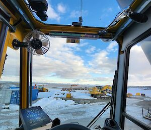 The inside of a yellow crane cabin in foreground, with boom extended and hook mid view, overlooking a snowy slope with a container and machinery.