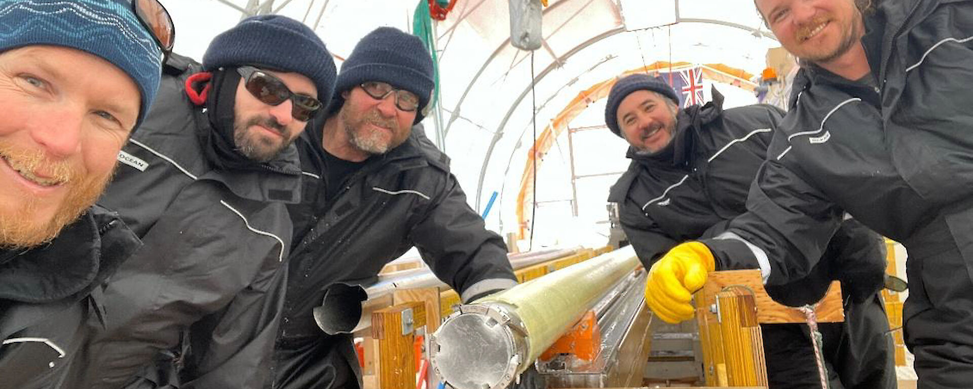 Five people crouched around the end of a ice core drill barrel containin ice that formed about 11,700 years ago.