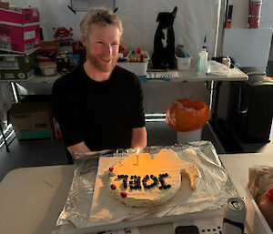 A man sitting at a table with a pavlova on a large square tray in front of him. The pavlova is shaped like Antarctica with his name, Joel, spelled out in blueberries and a candle at the site of the drilling at Dome C North.