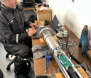 A man working on a computer set up on a wooden bench next to the electronics component of the ice core drill.