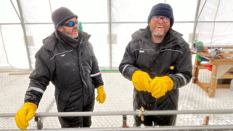 Two men wearing thick yellow gloves and drilling suits share a laugh in the ice core drilling tent.