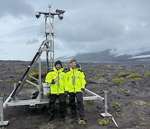 Two men in yellow stand in front of a large metal pyramid structure with a camera at the top