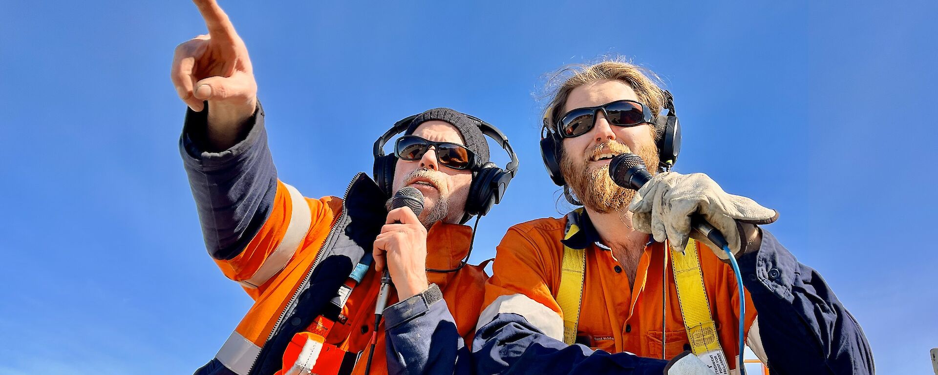 Two men in high visibility clothing look out over the camera, holding microphones and pointing into the distance.