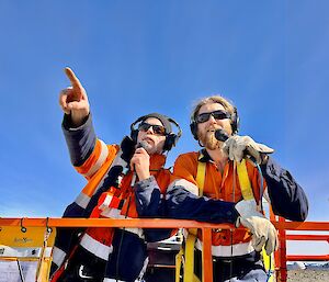 Two men in high visibility clothing look out over the camera, holding microphones and pointing into the distance.