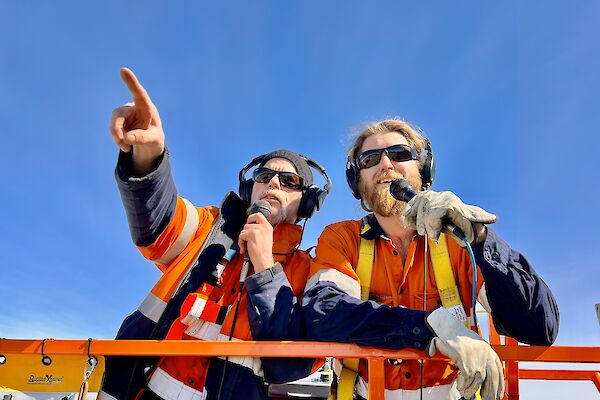 Two men in high visibility clothing look out over the camera, holding microphones and pointing into the distance.