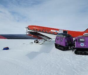 A purple hagglund positioned next to a red Basler aircraft on the snow.