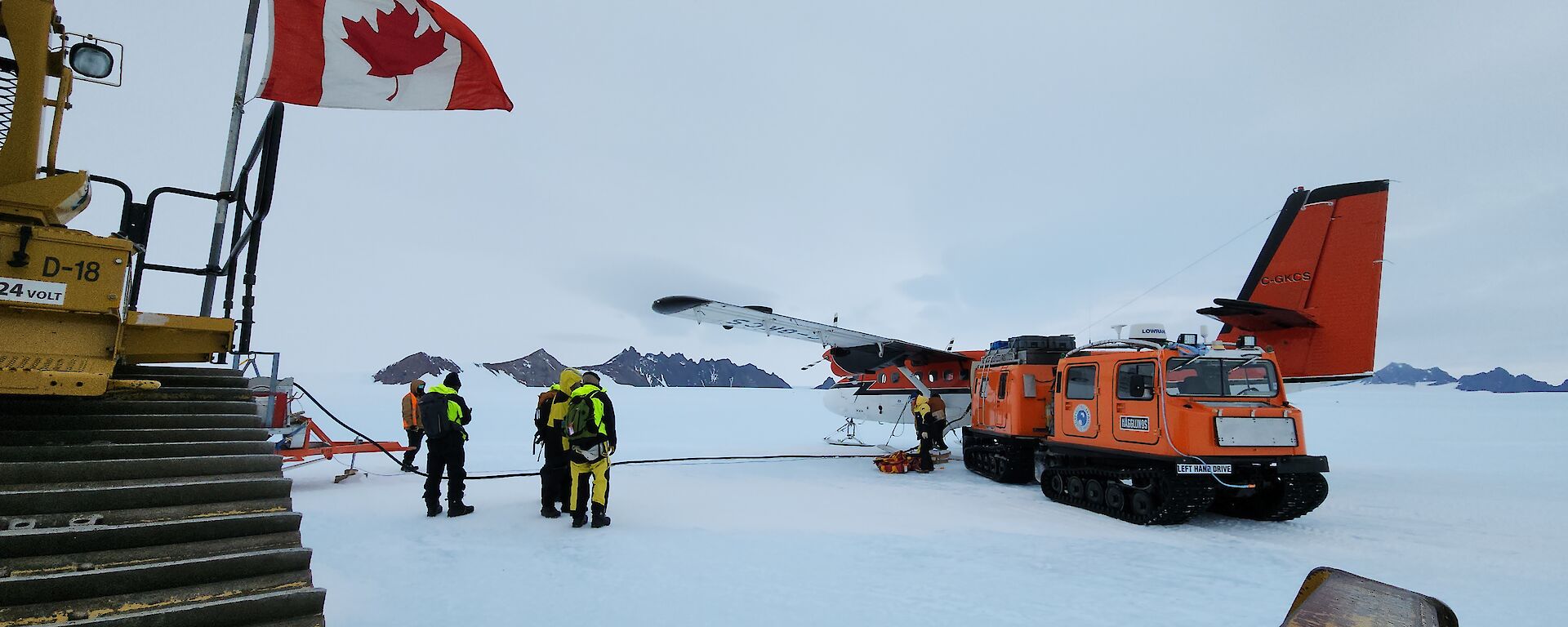 Four people standing near a Twin Otter and orange hagglund. Part of a tractor is visible to the left of the image with a Canadian flag flying.
