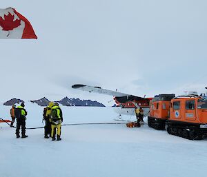 Four people standing near a Twin Otter and orange hagglund. Part of a tractor is visible to the left of the image with a Canadian flag flying.