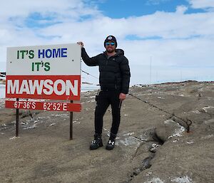 A person standing beside the Mawson station sign, which reads "It's home, It's Mawson".