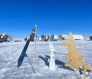 An Australian flag and a wooden cutout of a boxing Kangaroo next to a small snowman, with the ice drilling campsite in the background.