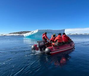 A red IRB with five people in it wearing red immersion suits in the foreground, in the back ground is an iceberg and blue water