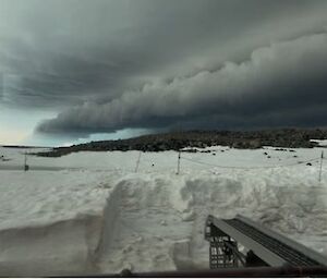 A cable tray and snow pile in the foreground, a large ominous dark grey cloud rolls across the sky in the background over a rocky hill on the horizon