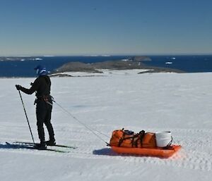A skier in the foreground with a sled attached at the waistline via a tow bridle, a snowy slope looking down to the water in the background