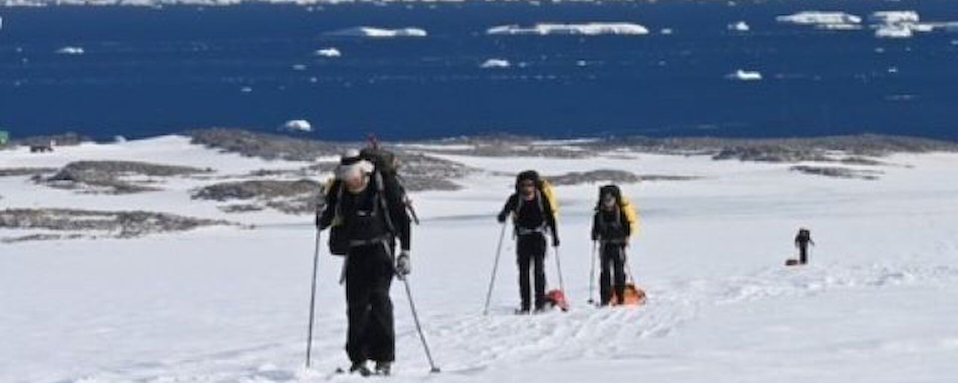 Three skiers in the foreground, two towing sleds, and a fourth skier in the distance also towing a sled, with views of the water