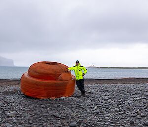 A massive orange ship fender, as tall as a person, with a man standing next to it on a grey stony beach