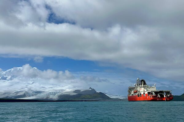 A red and white ship to the right, with clouds overhead and snow capped mountains in the distance