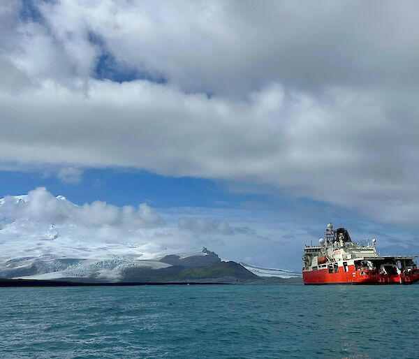 A red and white ship to the right, with clouds overhead and snow capped mountains in the distance