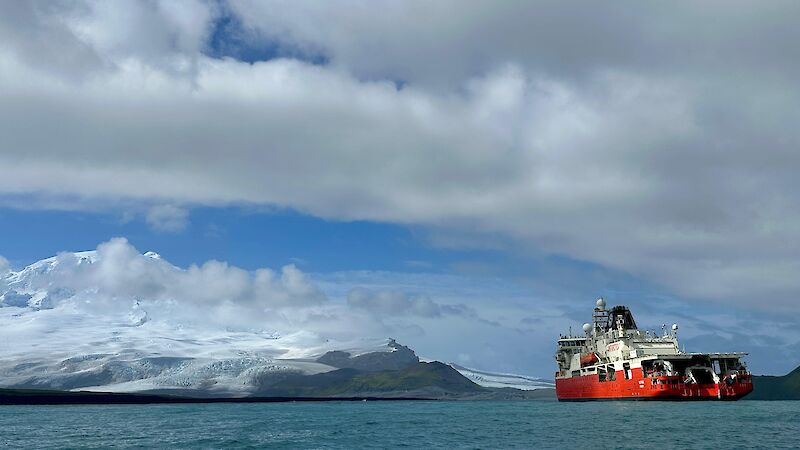 A red and white ship to the right, with clouds overhead and snow capped mountains in the distance