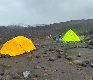 Two yellow tents on a grey rocky hillside with snow capped mountains in the background