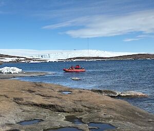 A small inflatable rubber boat pulling in to the rocky coastline, with an elevated snowy peak behind.