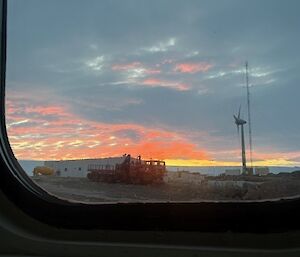 The view of a sunset, a station building, a truck, a wind turbine and a communications aerial, from a square window frame.