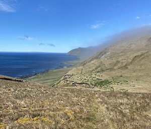 A grassy, hilly landscape abuts the edge of the blue ocean with blue cloudless skies above.