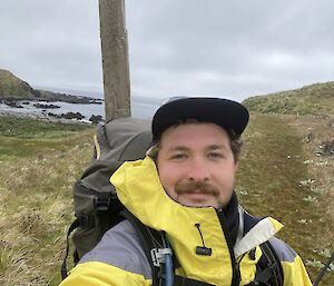 A smiling man wearing a yellow raincoat and a backpack stands in a grassy landscape with an ocean bay behind him.
