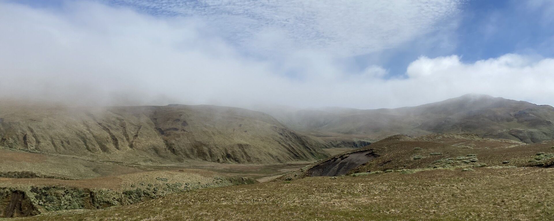A rugged range of hills rises out of a flat grassy landscape with a cloudy sky above it.