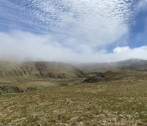 A rugged range of hills rises out of a flat grassy landscape with a cloudy sky above it.