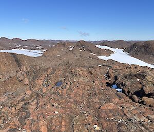 A view looking down over and expanse of medium sized grey/brown rocky hills and snow from a height, junder a blue sky.