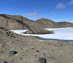 A rock, approximately the size of a car sits in a frozen lake, the edges of which are beginning to melt. The grey rocky hills in the background have dramatic black stripes on the.