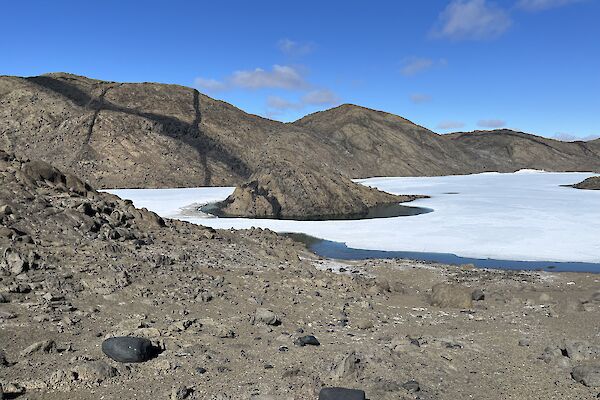 A rock, approximately the size of a car sits in a frozen lake, the edges of which are beginning to melt. The grey rocky hills in the background have dramatic black stripes on the.