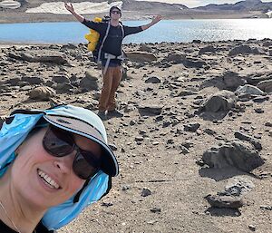 A man wearing a bright yellow pack stands with his arms raised in celebration in front of a sparkling lake. In the foreground a woman wearing a duck blue hat is smiling in a selfie
