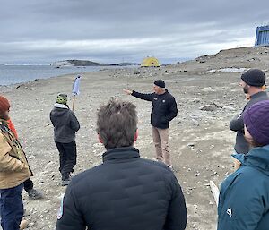 A group of people look on as a man wearing a black jacket and beanie points to something off screen