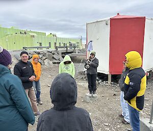 A group of expeditioners wearing warm jackets stand in a group in front of a small red and white building