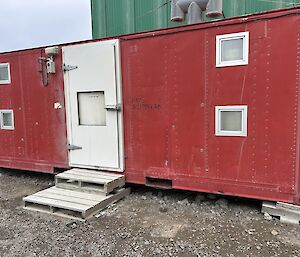 A red building, approximately the size of a shipping container with a white door and four windows, spaced out for bunk beds, two on each end of the container, either side of the door.
