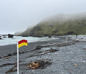Two red and yellow flags about 50 metres apart set into the sand on a misty, pebbly beach near the water's edge.