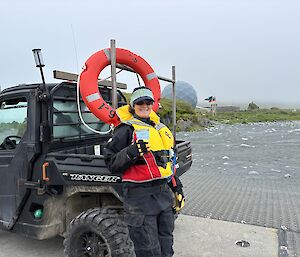 A smiling woman wearing a dry-suit and life jacket stands next to a black Polaris 4WD with an orange life ring on the back.