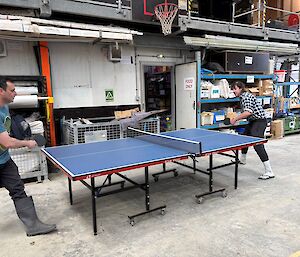 Two men play table tennis inside a warehouse.