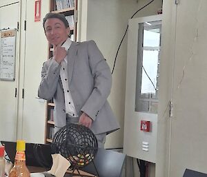 Bingo balls and bingo cards lay on a table in the foreground, Josh in the background donning a grey suit and pants, with powder in his hair