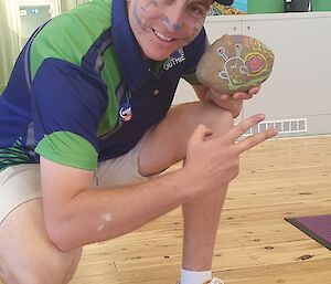 A photo of Josh knealing into the photo, holding up his rock art, the green walls and wooden floor of the youth centre in the background
