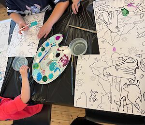 A photo taken from above a table, with childrens hands painting pages of Antarctic themed pictures