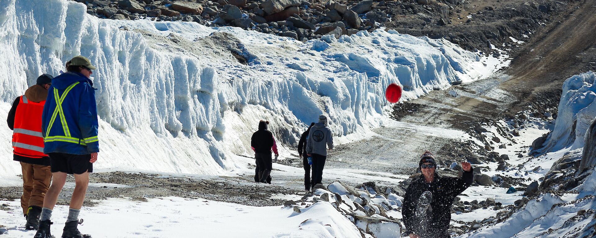 Three people in the foreground, one kicking a football. Three people in the background walking down Wharf road at Casey station, with snow surrounding the road half way down.