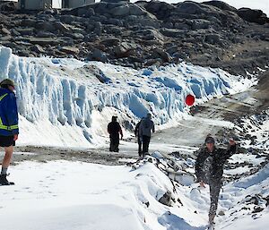 Three people in the foreground, one kicking a football. Three people in the background walking down Wharf road at Casey station, with snow surrounding the road half way down.