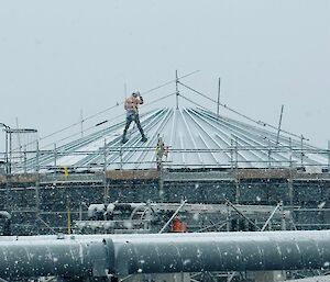A man wearing high visibility clothing and a harness stands on top of a metal frame, in snow