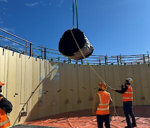 A large black bundle is lowered into a circular frame area, being guided by two people wearing high visibility clothing.