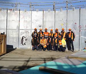 A group of 12 people wearing high visibility clothing pose for a photograph in two rows, next to artwork drawn on the wall behind them of a penguin that says 'Davis 79th is Fire'.