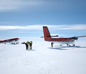 Two red and white aircraft on a skiway. Two people are walking towards one of the aircraft.