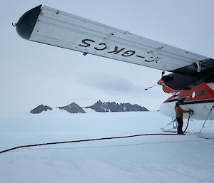 A man refuelling a twin otter aircraft, with a view of mountain peaks in the distance.