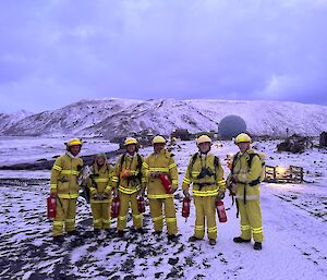Six expeditioners wearing yellow firefighting outfits stand in the snow in the pre-dawn light after responding to a fire alarm.